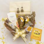 A beautifully arranged vishu gift basket for womens containing a bottle of banana chips, 4 Ferrero Rocher chocolates, Jimikki earrings, a greeting card, a Kerala saree, a personalized photo card, saffron and Sharkkaravaratty, and a bottle of Yardley Mist Perfume.